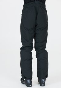 Zwarte snowpants met verstelbare banden, gestructureerde stof, volumineuze pasvorm en ritssluitingen bij de enkels. Voorzien van stevige zwarte skischoenen.