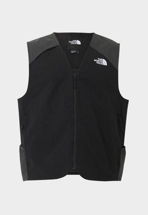 Mladý muž nosí černou vestu na zip přes černé tričko s krátkým rukávem s logy The North Face a hnědé kalhoty, stojící před bílým pozadím.