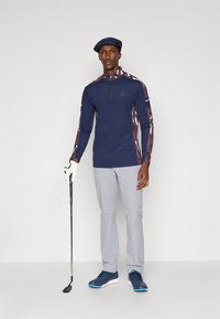 Navy long-sleeve golf shirt with patterned shoulders, gray pants, white glove, and blue shoes. Model holds a golf club.