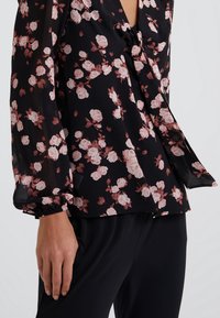 Blusa negra con estampado de flores rosadas, de tela transparente, con mangas largas abullonadas y cierre de lazo en el escote. Los pantalones negros son parcialmente visibles.