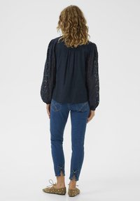 Blouse bleu marine avec manches en dentelle texturée, associée à un jean bleu orné de nœuds sur l'ourlet. Ballerines camel avec clous décoratifs.