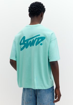 Osoba nosiaca voľné mentolovo zelené tričko s výrazným tyrkysovým grafickým nápisom na chrbte a modré rifľové šortky, stojaca na bielom jednoduchom pozadí.