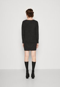 Osoba s krátkými vlasy, oblečená v šedých šatech a černých kozačkách po kolena, stojí zády k jednoduchému bílému pozadí.