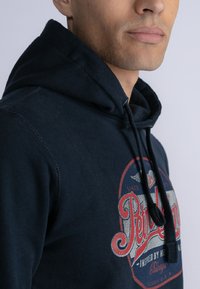 Marineblauwe hoodie met een vooraanstaand graphic, met de tekst "Peterson" in rood en wit. Het heeft strikkoorden en een zachte, gestructureerde stof zichtbaar.