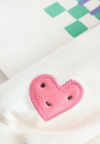 Haut en tissu blanc avec un cœur rose brodé souriant, bordé d'une couture rose, et des motifs géométriques verts et violets.