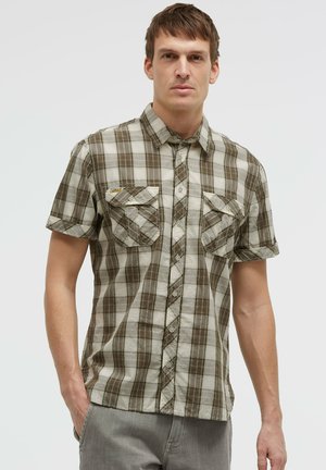 Mann trägt braun-beiges kariertes kurzärmeliges Hemd mit zwei Brusttaschen vorne, steht mit einer Hand in der Tasche.