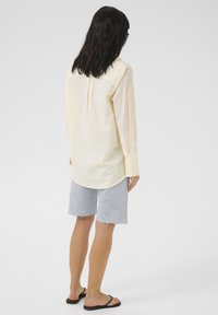 Camicia a maniche lunghe gialla chiara con orlo arrotondato e una pence dietro, abbinata a pantaloni shorts in denim blu con bordi sfilacciati. Infradito neri.