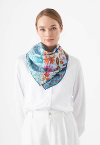 Seidenschal in Quadratform mit einem lebhaften floralen Muster in Pink und Blau, umrandet von geometrischen Designs, um den Hals über einer weißen Bluse getragen.