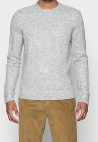 Uomo che indossa un maglione lavorato a maglia grigio chiaro e pantaloni in corduroy color senape, in piedi contro uno sfondo chiaro e semplice.