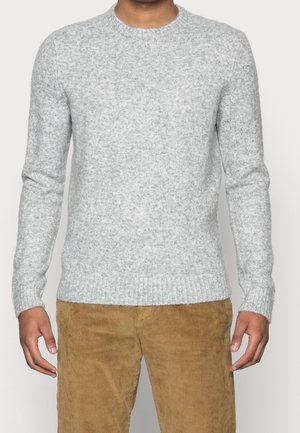 Uomo che indossa un maglione lavorato a maglia grigio chiaro e pantaloni in corduroy color senape, in piedi contro uno sfondo chiaro e semplice.