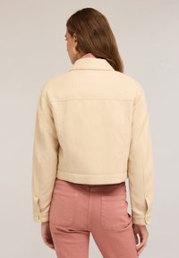 Bežová zkrácená bunda hladké textury s límečkem a knoflíkovými manžetami. V kombinaci s pastelově růžovými kalhotami.