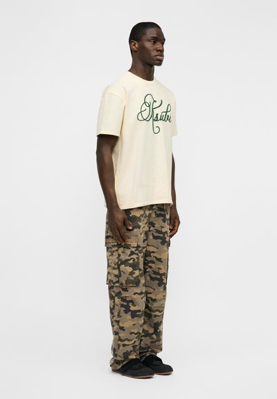 Κοντομάνικη κρεμ μπλούζα με πράσινα γραμμένα γράμματα, συνδυασμένη με παντελόνι καμουφλάζ cargo με πολλαπλές τσέπες. Μαύρα παπούτσια slip-on σε λευκό φόντο.