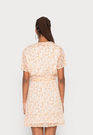 Femme aux longs cheveux tressés portant une robe fleurie claire à manches courtes avec un ourlet volanté, debout de dos sur un fond uni.