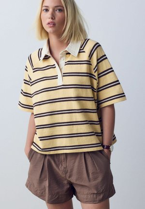 Junge Frau mit kurzen blonden Haaren, die ein gelb-braun gestreiftes Poloshirt und braune Shorts trägt, mit den Händen in den Taschen.