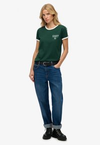 Groen T-shirt met korte mouwen en een witte kraag en manchetten, voorzien van de graphic "SDRY 82". Gekocht in combinatie met opgerolde blauwe spijkerbroek en zwarte laarzen.