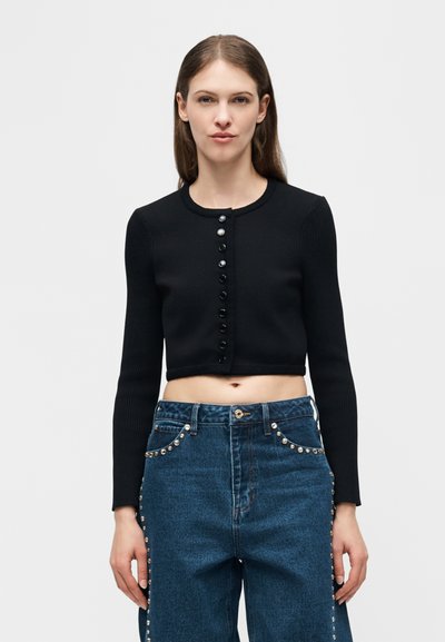 Νεαρή γυναίκα με μακριά καστανά μαλλιά που φοράει μαύρο κοντό πουλόβερ με κουμπιά και ψηλόμεσα μπλε τζιν με ασημένιες λεπτομέρειες με τρουκς.