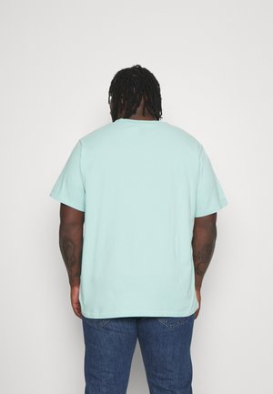 Man met dreadlocks die een lichtblauw T-shirt en blauwe spijkerbroek draagt, staat met zijn rug naar de camera tegen een effen witte achtergrond.