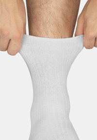 Weiße Baumwollsocke, die über ein Bein gezogen wird, mit gerippter Textur und eng sitzendem Schnitt. Der Stoff wirkt weich und dehnbar.