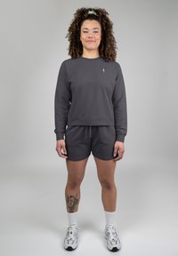 Felpa grigia con maniche lunghe, scollo rotondo e logo sul petto; pantaloni corti grigi abbinati; il modello indossa calze bianche e scarpe da ginnastica argento.