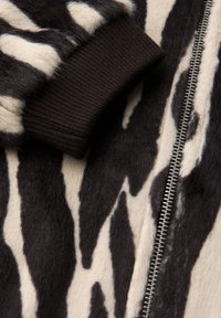 Veste bomber en fausse fourrure avec un motif zèbre noir et crème, des poignets en côte marron et des détails en zip argenté.