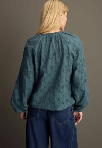 Blusa ricamata color turchese con motivi floreali, scollo arricciato e maniche lunghe. Indossata con jeans in denim scuri a vita alta.