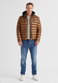 Braune Pufferjacke mit horizontalen Nähten, Reißverschlusstaschen und schwarzem Kapuze. Getragen über einem grauen Shirt, kombiniert mit blauen Jeans und weißen Sneakers.