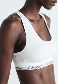 Weißer Baumwoll-Sport-BH mit rundem Ausschnitt und breiten Trägern, mit einem schwarzen 'Calvin Klein'-Logo auf einem weißen Band am unteren Rand.