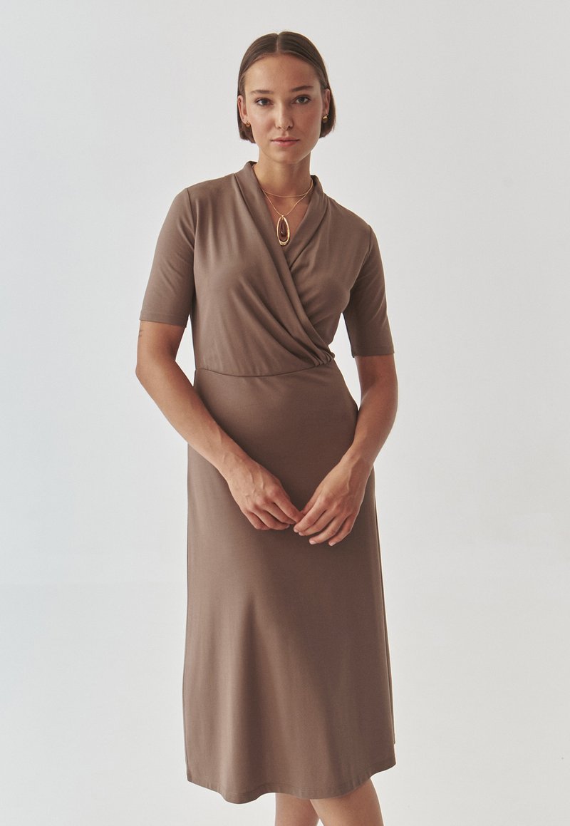 Vestido marrón envolvente con mangas cortas, confeccionado en una tela suave. Presenta un escote en forma de v y una silueta ajustada, que se extiende hasta la mitad de la pantorrilla.