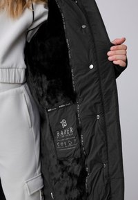 Zwarte jas met zwarte voering van zacht bont en drukknoopsluiting, met een label aan de binnenkant met de tekst "BAKER by Ted Baker".