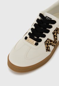 Adidași din piele albă cu șireturi negre și un panou lateral cu imprimeu leopard. Prezintă o placă cu logo de aur și o talpă din cauciuc maro texturată.