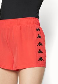 Pantalones cortos deportivos rojos con una cintura elástica, con logotipos negros de Kappa a lo largo del lateral. Tejido suave con un ajuste relajado y dobladillo recto.