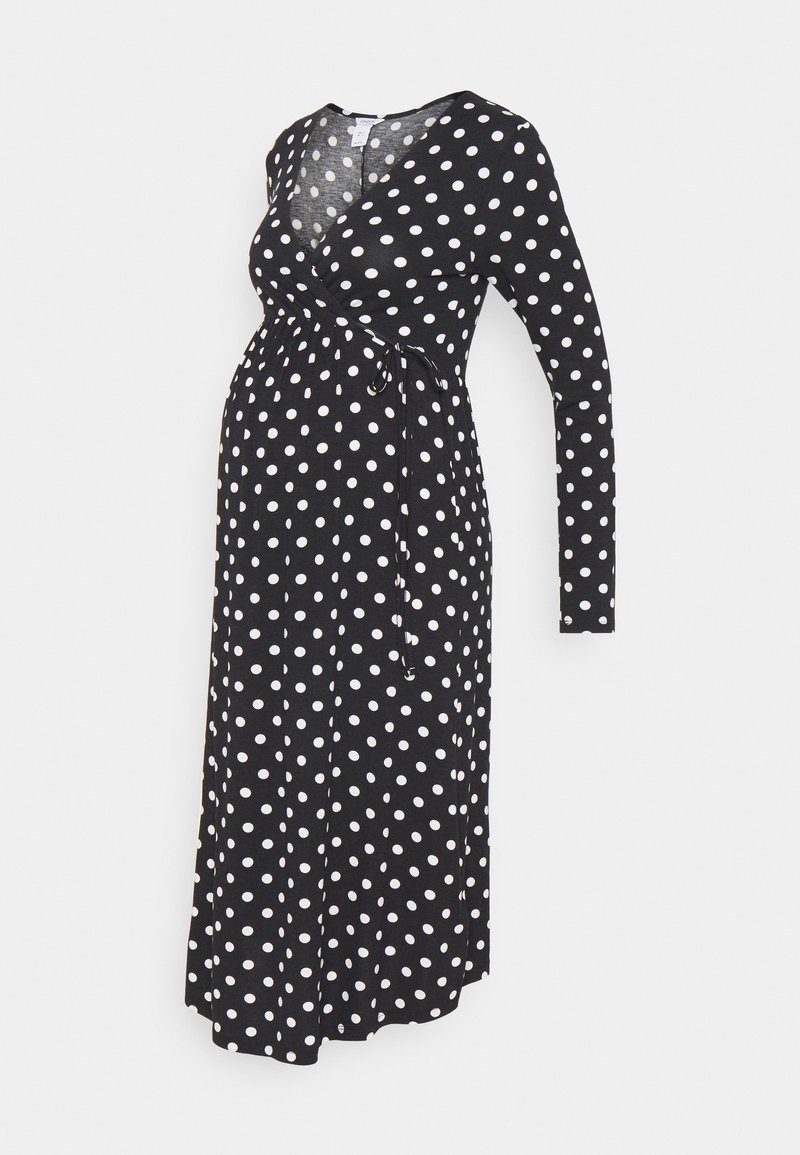 Robe de maternité noire à pois blancs, manches longues, style cache-cœur avec col en V et ceinture à nouer à la taille, exposée sur un mannequin.