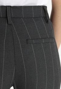 Tmavě šedé kalhoty s jemným proužkem, střih na míru, s poutky na opasek a zadními kapsami, nošené s bílým zastrčeným topem.