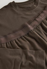 Brązowa bluzka z długim rękawem, z prążkowanym, okrągłym dekoltem, marszczonym detalem u dołu oraz teksturowaną opaską z napisem „SIGNATURE”.