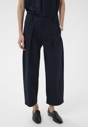 Személy laza, rövidített sötétkék nadrágot visel, amelyen ráncok vannak, fekete bebújós cipőkkel és egy ezüst karkötővel, fehér háttér előtt állva.