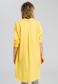 Robe chemise jaune en tissu léger, dotée d'un col, de manches boutonnées et d'un ourlet incurvé. Portée par-dessus des leggings à motifs.