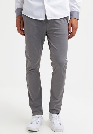 Mann trägt graue, eng anliegende Hose, weiße Turnschuhe und ein weißes, geknöpftes Hemd, steht mit locker an den Seiten hängenden Händen.