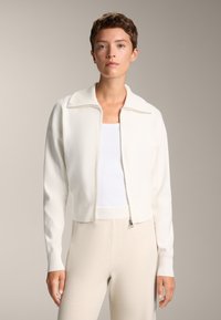Kurze, weiße Zip-Jacke aus strukturiertem Material mit breitem Kragen und gerippten Bündchen, getragen über einem weißen Tanktop und hellbeigen Hosen.