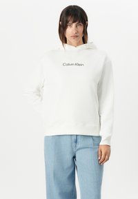 Felpa bianca in morbido tessuto con tasca a marsupio e logo nero "Calvin Klein" sul petto. Abbinata a jeans blu chiaro.