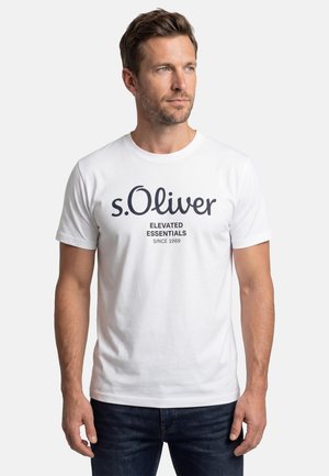 Mand med kort brunt hår iført en hvid t-shirt med teksten "s.Oliver Elevated Essentials Since 1969", stående foran en ensfarvet baggrund.
