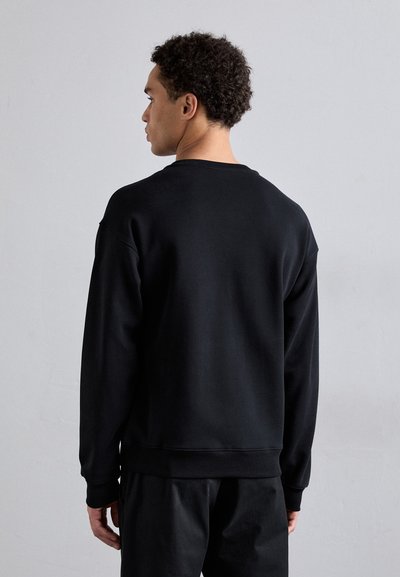 Schwarzer Pullover mit gerippten Bündchen und Saum, Rundhalsausschnitt und weicher Textur, getragen mit passenden schwarzen Hosen. Von hinten betrachtet.