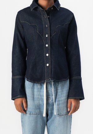 Veste en jean bleu foncé à boutons, avec empiècement style western et larges poignets, portée sur un jean en denim bleu clair avec taille à cordon.