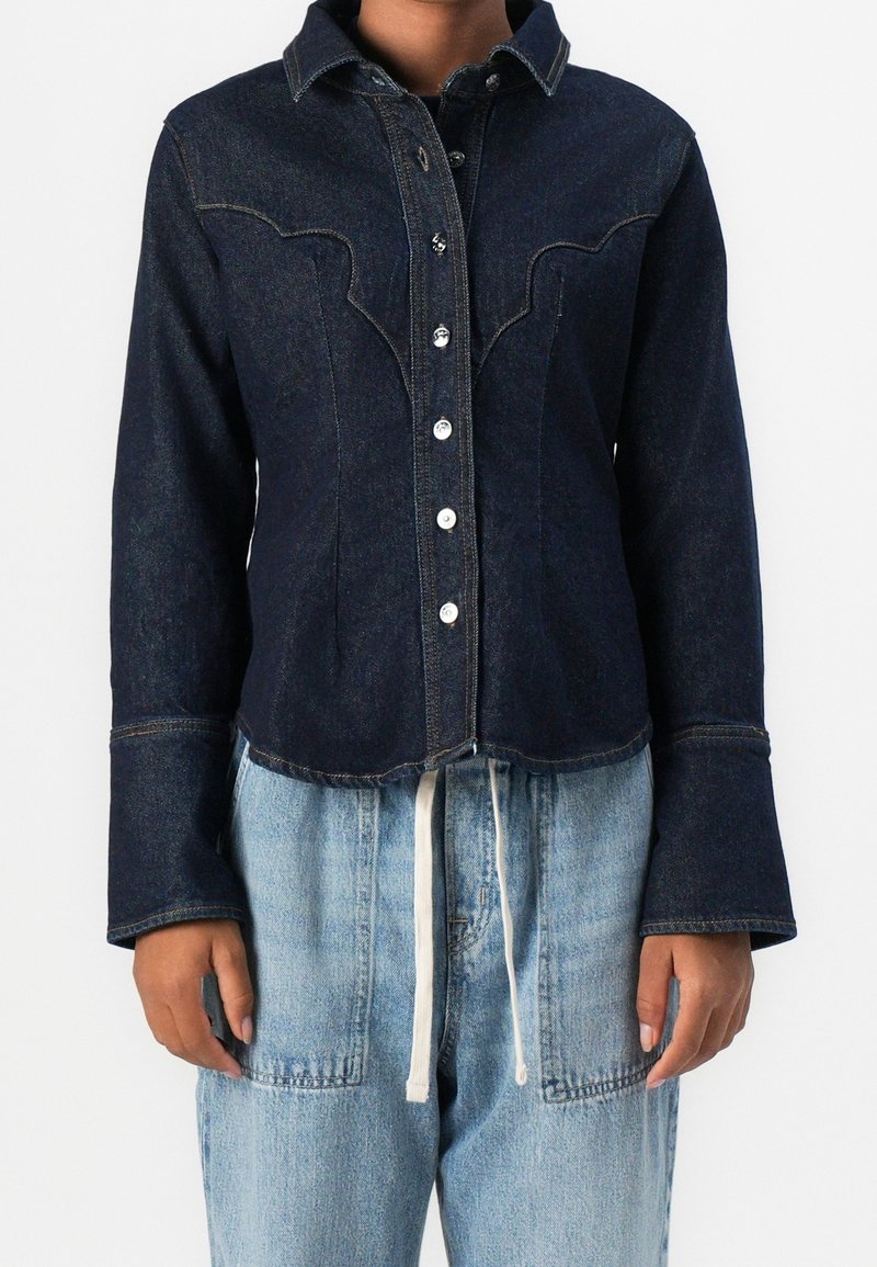 Veste en jean bleu foncé à boutons, avec empiècement style western et larges poignets, portée sur un jean en denim bleu clair avec taille à cordon.