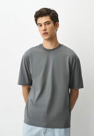 Junger Mann mit kurzem dunklem Haar, der ein schlichtes graues T-Shirt mit kurzen Ärmeln und hellblaue Jeans trägt, steht vor einem weißen Hintergrund.