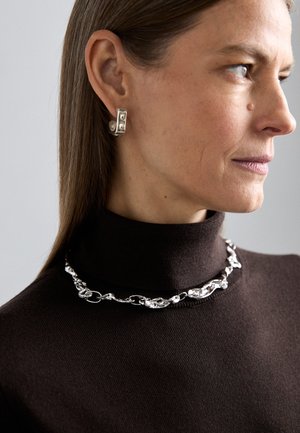 Ασημένια αλυσίδα λαιμού με οργανικούς κρίκους, φορεμένη πάνω από έναν σκούρο καφέ πουλόβερ με ψηλό λαιμό; αναδεικνύει γεωμετρικό ασημένιο σκουλαρίκι με κυκλικές λεπτομέρειες.