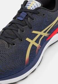 Detailní záběr na tmavě modrou běžeckou obuv se zlatým a červeným logem Asics, černými tkaničkami, horní částí z prodyšné textilie a bílou podrážkou označenou "FF Blast".
