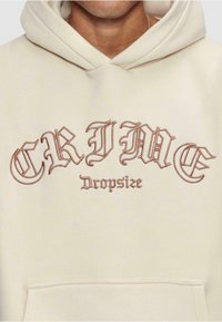 Haut à capuche couleur crème avec un grand texte gothique marron "CRIME" et "Dropsize" d'une texture douce et une poche kangourou à l'avant.