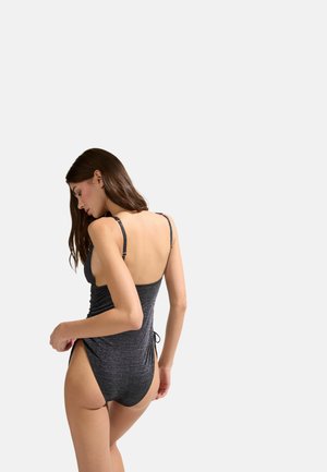 Femme portant un maillot de bain une pièce gris foncé scintillant avec de fines bretelles, posant de profil sur un fond blanc uni.