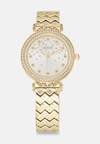 Gouden horloge met een gestructureerde armband, ronde wijzerplaat, witte wijzerplaat, diamantachtige accenten en gouden uurmarkeringen. Merklogo "GUESS" zichtbaar.