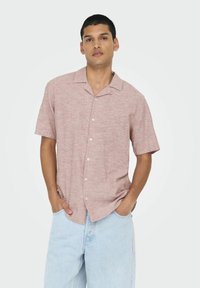 Camisa de manga corta con cuello en lino color rosa claro con un acabado texturizado, que presenta cinco botones frontales y un corte relajado, combinada con pantalones de mezclilla claros.